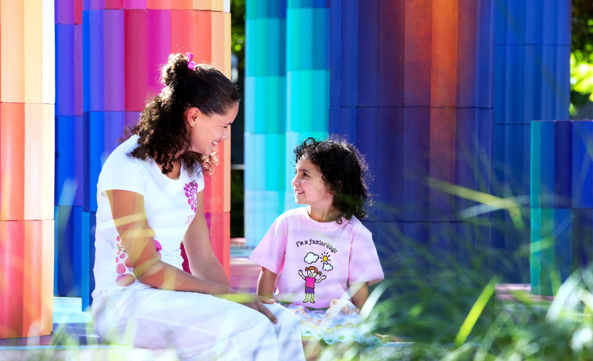 A big and little girl talk together in a colourful setting